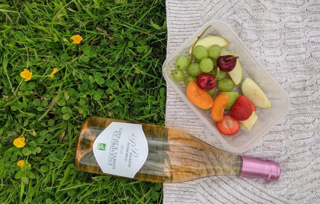 A bottle of rosé wine is balanced on a cream jumper laid out on green grass, next to fruit salad in a plastic tub
